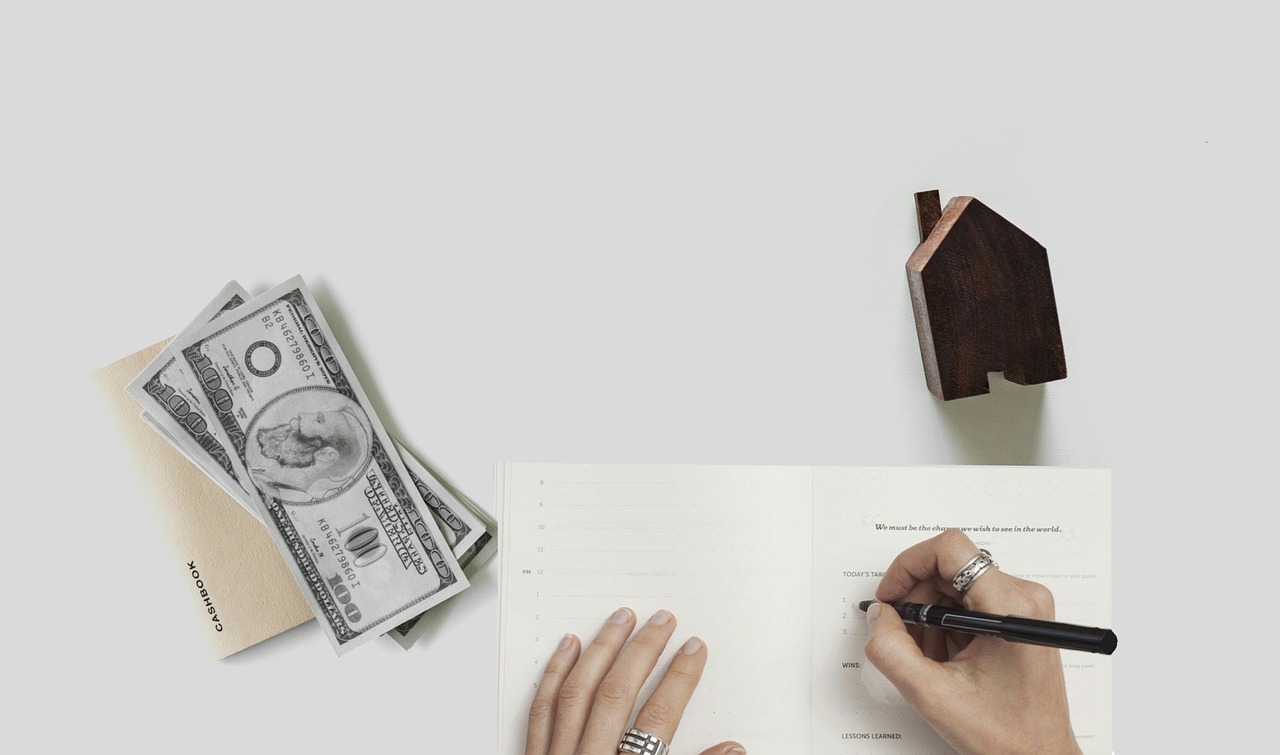 Hands writing in a planner beside cash and a wooden house, illustrating mortgage and estate planning.